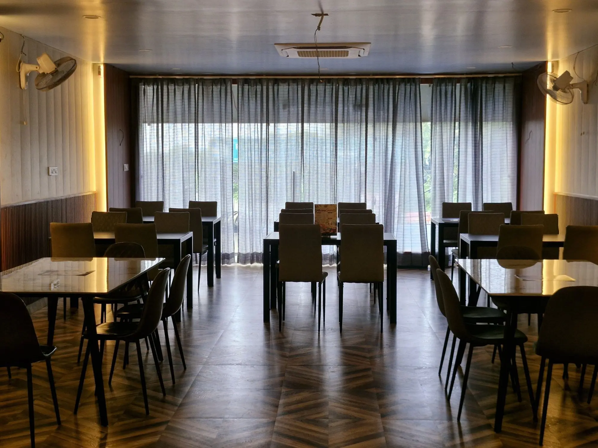 The curtained dining room: A calmer room: chevron floors, padded cream walls with warm edge lighting, and sheer curtains across the full-width glass.